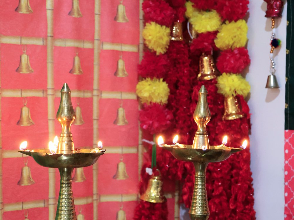 Two lit brass lamps against a red festival backdrop hung with marigold and bells